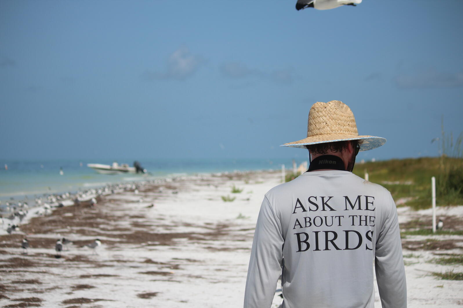 Evan standing away from the camera, with his "ask me about the birds" shirt in full view. He is standing on a beach, with water in the background.