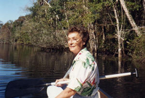 In this old photograph, a woman sits in a canoe and holds an oar across her lap. Her head is turned over her shoulder to face the photographer, who is sitting behind her.