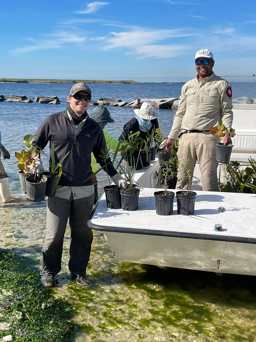 Two men hold up plants, with a boat and water in the background.