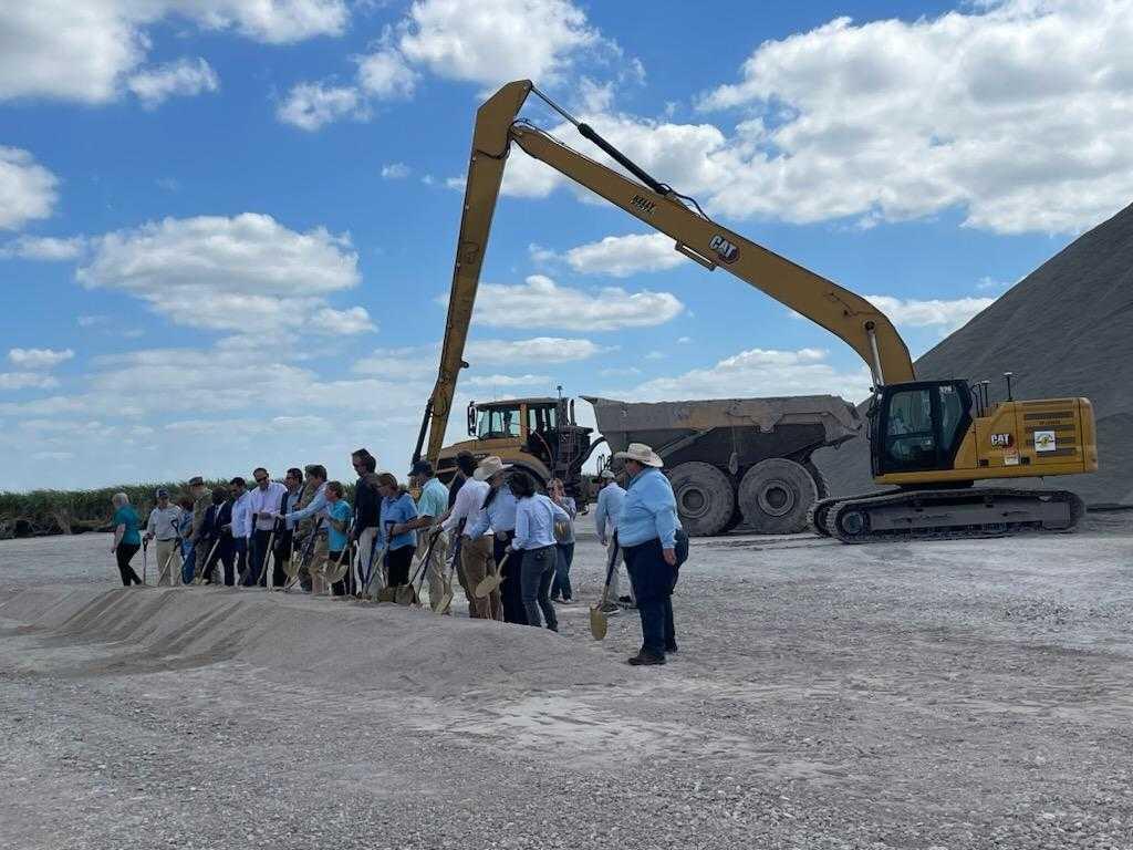 A group of people pose for a photo holding shovels, with an excavator in the background.