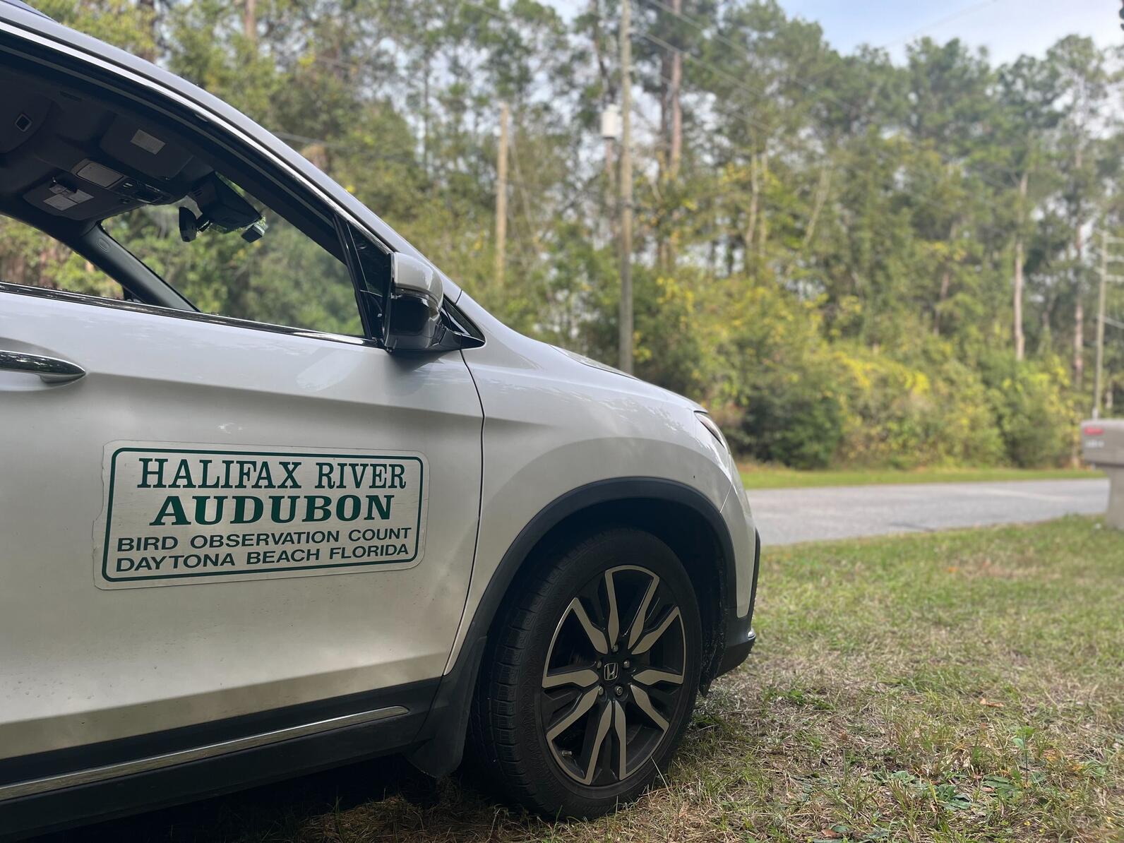 The front passenger side of a white SUV with a sticker reading "Halifax River Audubon - Bird Observation Count - Daytona Beach, Florda." The car is parked on the grass beside a narrow road through a wooded area.