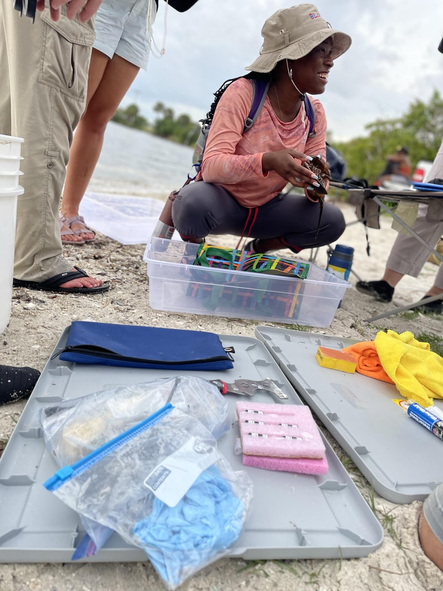 A woman crouches on a beach, with a plastic bin in front of her and additional equipment on the ground.