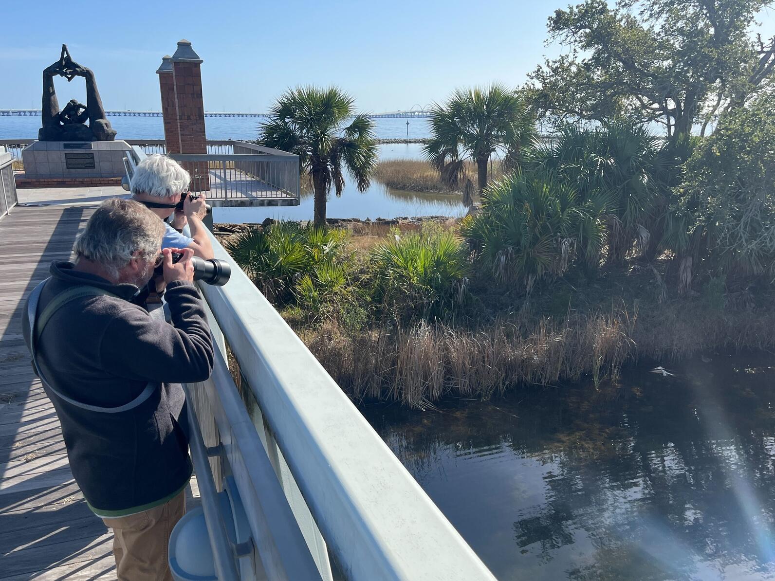 Two people stand on a bridge looking over the edge with binoculars at a small stream below.