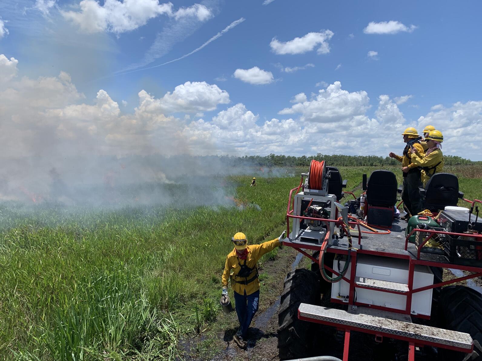 A person in yellow protective gear stands in front of a field with smoke in the near distance. Beside the person, a trio of other people stand atop a swamp buggy.