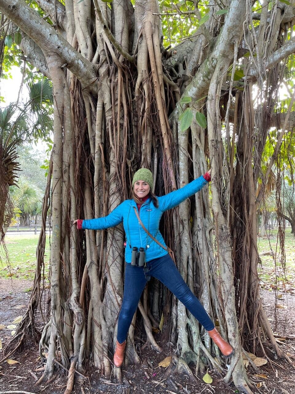 A woman stands with arms and legs outstretched on the roots of a large tree.