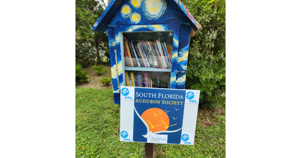 A Little Free Library, filled with books, with a South Florida Audubon Society sign hanging from the bottom.