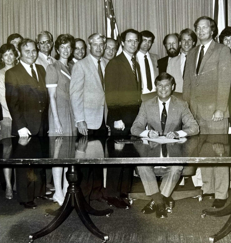 Black-and-white photo of a man sitting at a desk signing a paper with a group of people behind him.