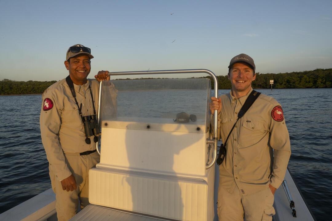 Two men smile at the steering wheel of a boat.