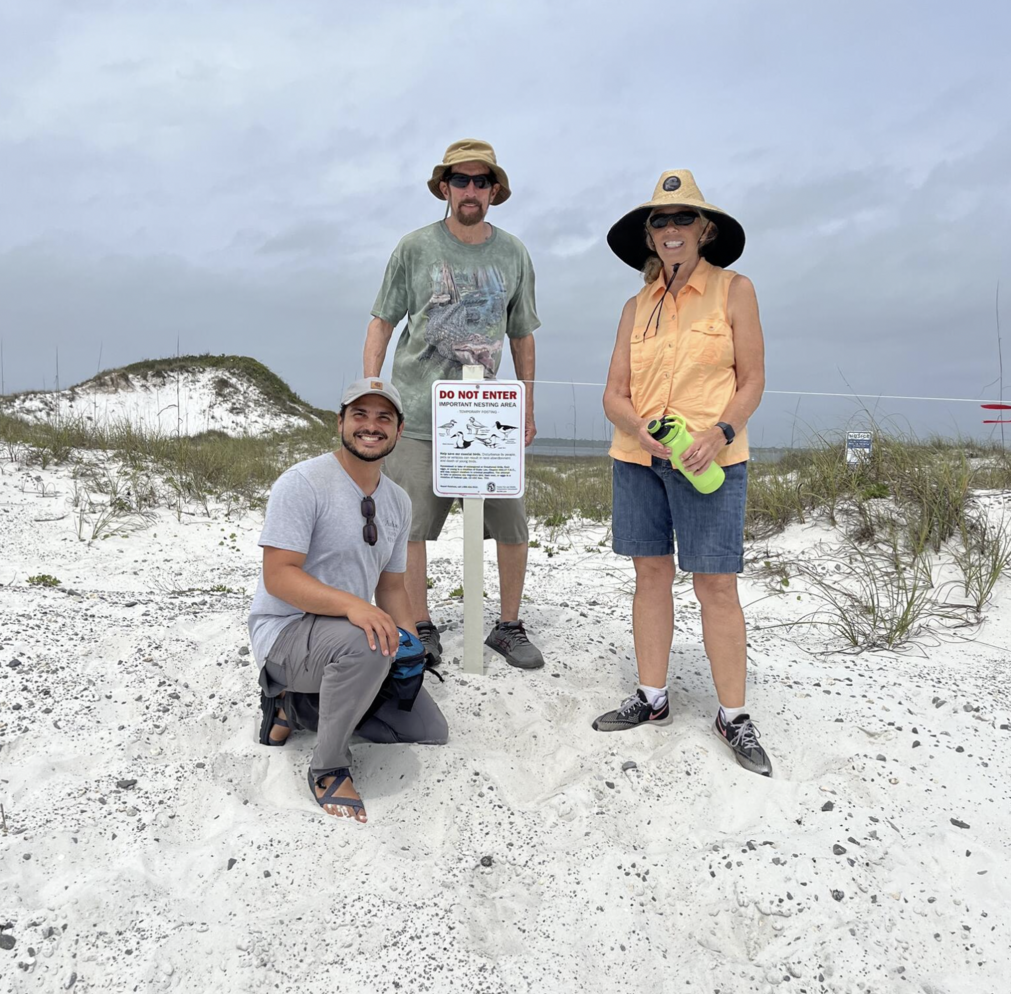 Three people stand on the sand and smile at the camera.